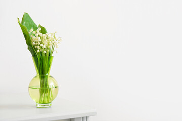 Vase with lily-of-the-valley flowers on shelf near light wall