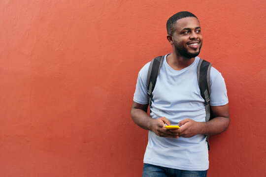 African Man with backpack and mobile phone smiling while leaning on wall