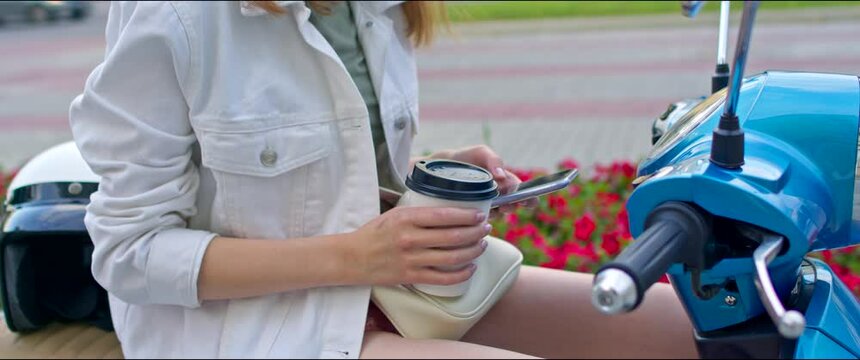 Young woman sits on a retro scooter. The girl holds a paper cup with coffee in her hand and uses a smartphone. Urban style.