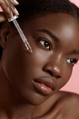 Portrait close up of beautiful black girl dropping serum collagen moisturizer on face. Serious young woman. Concept of face skin care. Isolated on pink background. Studio shoot - obrazy, fototapety, plakaty