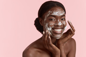 Close up portrait of beautiful black girl wash her face with cleansing face foam. Happy young woman looking at camera. Concept of face skin care. Isolated on pink background. Studio shoot - Powered by Adobe