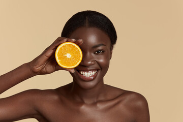 Portrait close up of beautiful african girl hold slice of orange. Happy young woman looking at camera. Concept of skincare. Isolated on beige background. Studio shoot