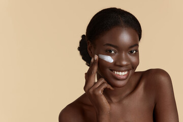 Portrait close up of beautiful black girl apply cosmetic cream on face. Smiling young woman looking...