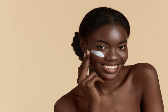 Portrait Close Up Of Beautiful Black Girl Apply Cosmetic Cream On Face. Smiling Young Woman Looking At Camera. Concept Of Face Skin Care. Isolated On Beige Background. Studio Shoot