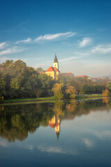 Scenic view of Zupnija Maribor Sv. Jozef church in misty morning landscape with colorful autumn trees, lush foliage, calm water of Drava river and reflection, blue sky with clouds, Maribor, Slovenia - obrazy, fototapety, plakaty