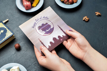 Female hands with greeting card for Eid al-Adha (Feast of the Sacrifice), Turkish sweets and Quran on dark background