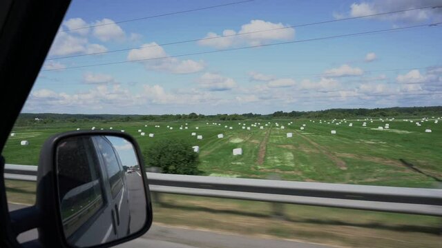 Round bales with silage wrapped in white plastic film on agriculture field view from car window