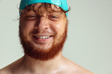 Close-up image of red-headed man in swimming hat posing isolated on gray studio background. Concept of sport, humor and body positive.