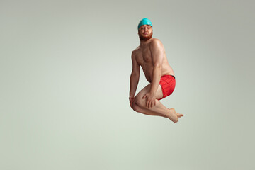 Funny cheerful swimmer. Cute red-headed man in red swimming shorts posing isolated on gray studio...