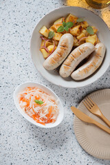 Fried german white sausages with potato salad and sauerkraut, vertical shot on a beige granite background, view from above