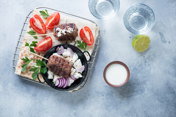 Grilled kofta with feta cheese, red tomatoes, white yogurt and flatbread, above view on a light-blue stone background, studio shot