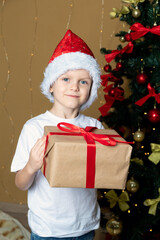 A boy in a New Year's hat with a smile holds a gift box with a bow in his hands. There is a Christmas tree behind. Selective focus.