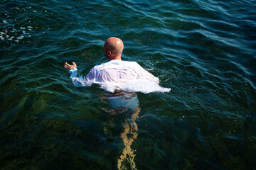 Adult man with shaved head in the ocean or sea in a wet white shirt