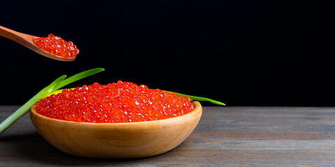 Red caviar in a wooden cup on a black background with a spoon and green onions. A large pile of bright caviar. Fresh delicious caviar. A place for advertising, logo, label, layout, mockup.