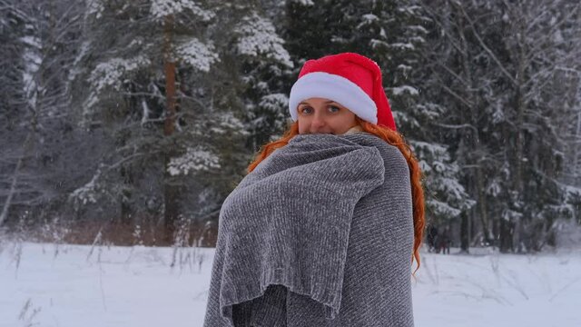 A woman in a red Christmas hat wraps herself in a blanket in a snow-covered park looks at the camera