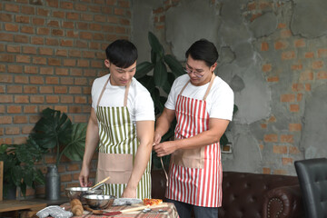 Happy Asian young LGBT gay couple making pizza together. A man helping his boyfriend put on an...