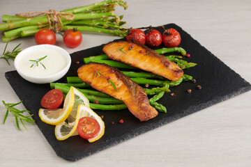 Delicious seasonal green asparagus and sliced smoked salmon on a rustic plate with lemon, salt, pepper, of lemon on a slate. Baked salmon garnished with asparagus and tomatoes with herbs.