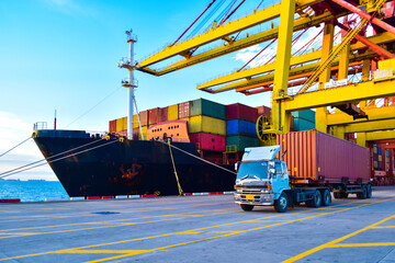 Truck carrying forty-foot container leaving port terminal with ship and quay crane on the...