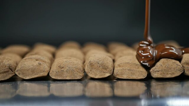 thick milk chocolate is poured onto brown candies in a row lying on culinary foil. Close-up, slow motion, dolly shot