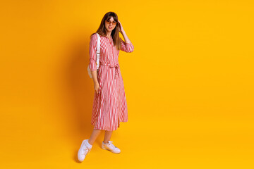 Indoor portrait of stylish woman wearing striped red dress, beige sunglasses and white bag, fashionable mood, studio yellow background.