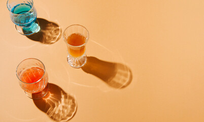 Creative sunlit layout made with glasses filled with colorful drinks on a pastel orange background. Summer day shadow ambient.