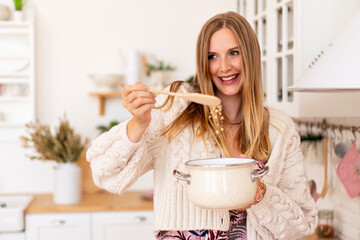 Pretty cute blonde house wife woman preparing food on her pink cozy stylish kitchen, wearing soft cream cardigan, holding pot with a tasty dish.