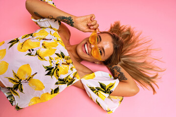 Positive summer studio portrait of cheerful blonde woman laying on the floor wearing trendy sunglasses smallish and laughing, trendy blogger outfit, pink background.