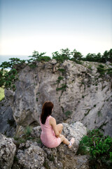 a girl in a striped pink dress sits on the edge of a cliff in the mountains