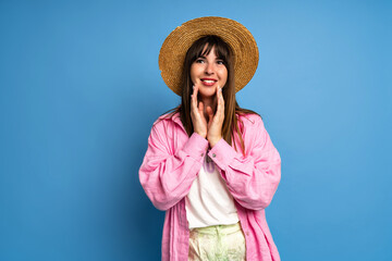 Summer portrait of pretty brunette woman wearing straw hat and bright linen shirt posing on studio blue background. Vacation style.