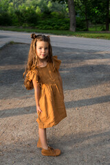 Selective focus portrait of beautiful smiling little girl with long dirty-blond hair wearing a tan linen dress and boots seen standing in park alley in rays of golden sun 	
