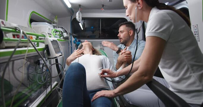 Caucasian young handsome husband supporting pregnant wife in ambulance car during labor. Man breathing and holding woman hand when in childbirth. On way to hospital with female doctor.