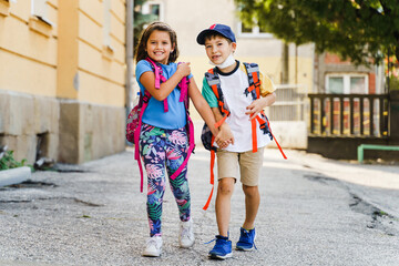 Front view full length portrait of small caucasian boy and girl brother and sister or little friends children going to school first grade pupils with backpack bags on their back in sunny day outdoor