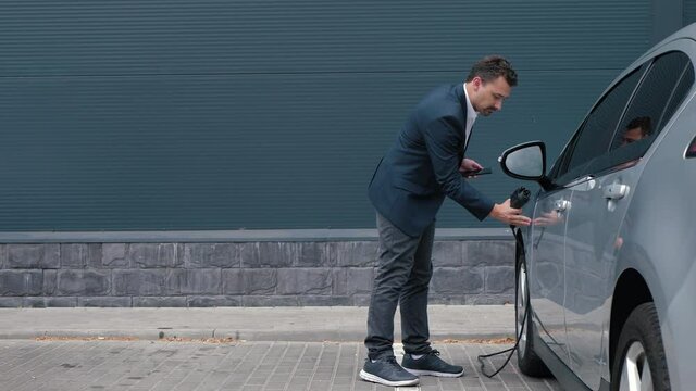 Businessman coming to electric car and plugging in charging cable. Man in jacket inserts power connector into EV car and charges batteries in parking lot, uses phone, activates switch on and leaves