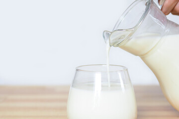hand pours milk from a jug into a glass on a wooden table on a gray background..