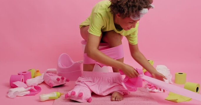Curly haired woman sits on toilet bowl rolls out paper dressed in domestic clothes spends morning time in restroom reveals herself defecates isolated over pink background. Human necessities.