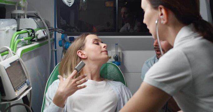 Caucasian beautiful woman in pain breathing hard and pushing when in labor and handsome husband supporting her while holding hand. Midwife in stethoscope listening heartbeat and helping in ambulance.
