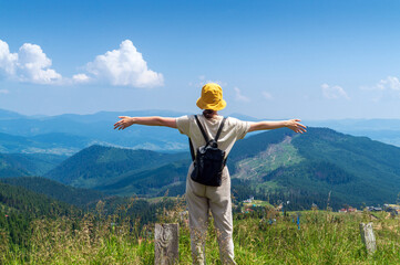 Girl traveler with a backpack stands with her arms outstretched in the mountains in summer. The top of the mountain evokes good or uplifting mood