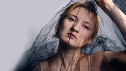 Fashion Model with a black net veil. Isolated woman under black cloth Net. Portrait of a blonde girl with a serious facial expression trying to escape. High-quality studio shot.