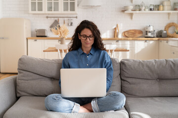 Millennial woman in eyeglasses use laptop computer sitting on couch in living room. Young...