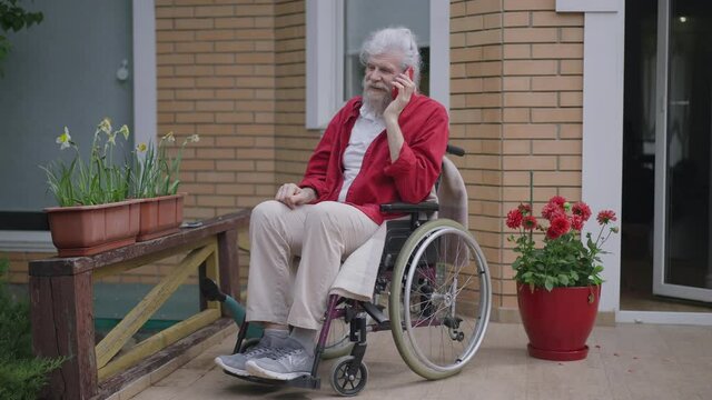 Smiling Happy Senior Disabled Man In Wheelchair Talking On The Phone Outdoors On Porch. Wide Shot Of Positive Cheerful Caucasian Retiree Sharing With Friend Or Family Chatting On Smartphone