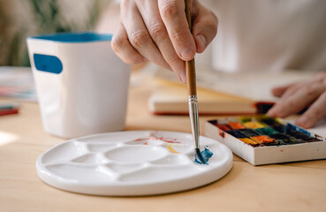 A young artist paints with watercolors in a notebook sitting at a wooden table. Beautiful male hands are holding a brush. Close-up. Sun rays and pleasant atmosphere. craft concept.The artist in action
