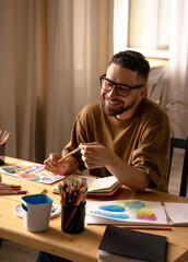 Portrait of dark haired smiling caucasian white male artist with a beard in a glasses, khaki t-shirt in studio, sunlight. Colored pencils and paintbrushes on a wooden table. Lifestyle. Real emotions