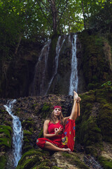 Naklejka na ściany i meble Front view portrait of adult caucasian woman 30 years old sitting in nature in front of waterfall in red dress stretching in yoga pose asana - Spirituality inner peace and relaxation concept