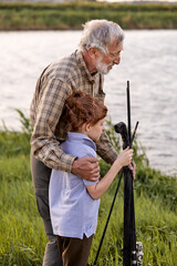 Nice family fishing together from riverside in the evening, pleasant grandfather and grandson in nature together, looking at side, senior male in checekered shirt helping, supporting child boy, teach