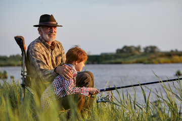 Side view portrait of man and little grandson sitting near river and fishing with fishing rods, spending nice good time together, copy space. in nature, in countryside - Powered by Adobe