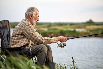 Senior man fishing outside in evening on lake in summer sitting on chair, enjoy spending time in...