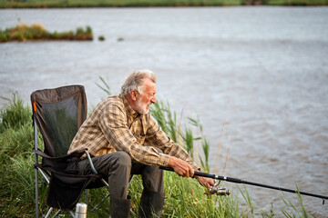 Fishing is very common hobby in coastal areas areas with rivers. Senior caucasian Man fishing in lake alone, in countryside, holding Fishing rod in hands, in casual wear
