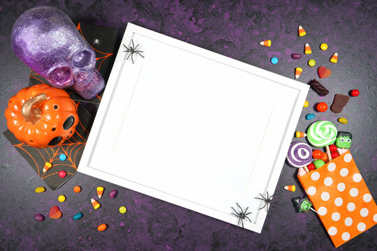 Halloween theme white wood wall art frame mockup flatlay on textured purple background with purple and orange skull, pumpkin and trick or treat candy. Product mock up with negative copy space.