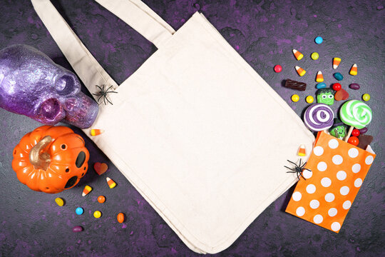 Halloween Theme Tote Bag Mockup Flatlay On Textured Purple Background With Purple And Orange Skull, Pumpkin And Trick Or Treat Candy. Product Mock Up With Negative Copy Space.