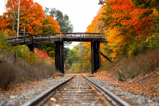 An old wooden bridge sits over railroad tracks surrounded by orange and yellow fall color trees in autumn_02